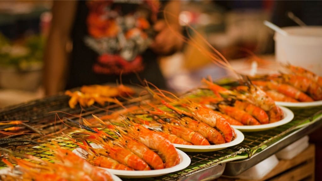 Plates of grilled prawns are arranged in a row on a wire rack at a food market stall.